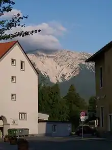 Une montagne, se faisant recouvrir d'une nappe de nuages à l'arrière plan, est limitée par des habitations et de la végétation au premier plan.