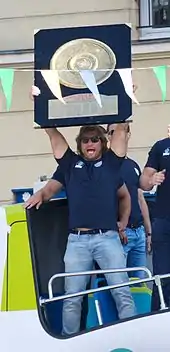 Homme en tenue de ville, en pied, tenant au dessus une planche représentant le bouclier de Brennus.