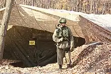 Soldat en uniforme dans une forêt. Son gilet tactique et son casque sont dans un motif de camouflage vert tandis que le reste de sa tenue est dans un motif camouflage beige.