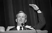 Photographie en noir et blanc d'un homme en costume et faisant un discours devant des micros