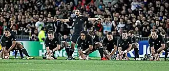 De face, plusieurs hommes, en tenue de rugby noire, accroupis. Un autre joueur les bras étendus sur le côté, est au centre, au milieu des des autres.