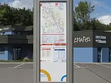 Photographie en couleurs d’un panneau gris et bleu devant un cabinet vétérinaire, qui marque un arrêt de bus. Il comporte les fiches horaires de passage de deux lignes : une du réseau de Rumilly et une du réseau d'Annecy.
