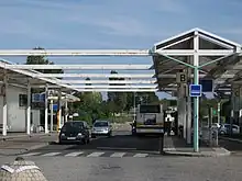 La photographie présente un centre bus en été. Un bus de la SEMTAO stationne sur le quai B, une personne descend du bus. Des voitures circulent sur le quai A. Le quai C est vide. Les préaux des quais sont soutenus par des piliers métalliques peints en blanc.