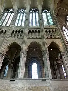 Trois piliers sont visibles reliés par un mur de pierre. Au-dessus, le triforium et quatre fenêtres hautes de verre blanc.