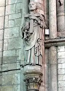 Statue de calcaire blanc représentant un homme barbu portant une pierre et une croix.