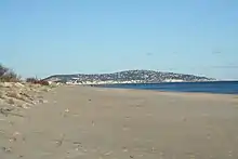 Sable au premier plan. En arrière-plan, on devine des immeubles au ras de l'eau et des maisons éparpillées sur une hauteur.