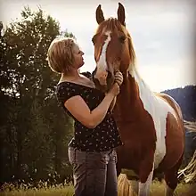 Photographie en couleur d'un cheval de couleur rousse et blanche et d'une femme qui lui touche la tête.