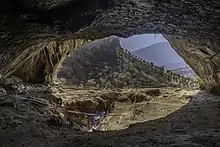 Photographie de l'entrée d'une grotte vue de l'intérieur. À l'extérieur on peut voir le versant d'une autre colline et une plaine plus en arrière. À l'intérieur, on peut y voir un grand trou creusé par les archéologues entouré de barrières de sécurité.