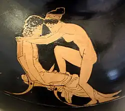 Photographie en couleurs du détail de l'ornementation d'une céramique à figures rouges sur fond noir, représentant une scène d'accouplement entre un homme assis et une femme nue, portant un bandeau dans ses cheveux.