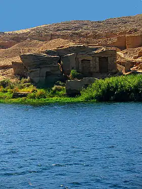 Vue d'un paysage égyptien, de l'eau au premier plan, des grottes et une montagne aride au second plan