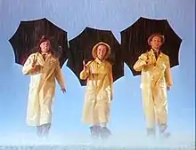 Image du film où deux hommes et une femme souriants, vêtus de longs imperméables jaunes et portant des parapluies noirs, marchent de concert sous une pluie battante en direction de la caméra.