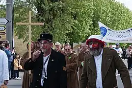 Un homme barbu porte un bonnet de nuit et a le visage recouvert de maquillage rouge. À sa droite, un homme déguisé en gendarme l'accompagne.