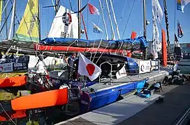Vu de trois quarts arrière, le bateau bleu et gris, safrans orangés relevés, amarré au ponton.