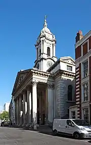 Photographie couleur. Bâtiment vu en enfilade sur le côté droit, précédé d'un portique à 6 colonnes et fronton néo-classique, surmonté d'un clocher carré à toit bulbeux, avec des horloges sur chaque pan.