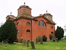 Photo d'un bâtiment en briques rouges aux murs imposants, situé au milieu d'une pelouse jonchée de pierre tombales