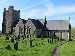 Photo d'un bâtiment allongé aux murs en pierres grises et au toit en ardoises, flanqué d'une grande tour carrée sur la gauche, au milieu d'une pelouse jonchée de pierres tombales
