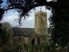 Photo d'un bâtiment allongé aux murs gris et au toit en ardoises, avec une tour carrée trapue en son centre. La silhouette d'un arbre au premier plan encadre le sujet