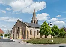 Vue de l'église : on voit le portail et la nef de l'église, et derrière apparait le sommet du clocher en flèche. Trois arbres bordent l'édifice, des pots de fleurs sont positionnés devant le portail.