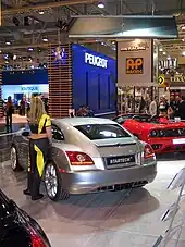 Une voiture 2 portes grise vue de l'arrière gauche exposée dans un salon aux côtés d'autres automobiles. Une femme s'appuie sur la portière conducteur de la voiture.