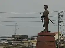 Vue de loin d'une statue en ciment montrant un homme moustachu tenant un fusil dans une main et un bâton dans l'autre, portant des vêtements traditionnels. Il est debout sur un piédestal de pierre polie.