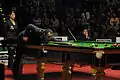 Stephen Maguire, jouant en extension, avec une jambe appuyée contre la table.
