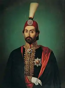 Portrait du sultan ottoman Abdülmecid Ier à la barbe courte portant un fez surmonté d'un plumeau, un manteau noir doublé de rouge et une veste aux broderies dorées sur laquelle se trouvent plusieurs médailles en forme d'étoiles.