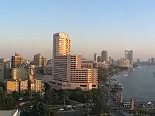 High-rises, including a multi-story white building in the foreground, dominate a sunset view of Cairo alongside the Nile, which is shown flowing under a bridge carrying a busy street