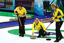 Portrait de trois joueuses de curling en veste jaune et pantalon noir, l'une accroupie au centre tenant une pierre à la main, les deux autres tenant un balai.