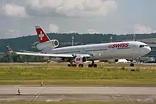 Photo couleur d'un avion de ligne à trois réacteurs, dont deux de chaque côté des ailes et l'autre sur la queue. L'avion, aux couleurs blanches et rouges avec une croix blanche sur fond rouge sur la queue, est au décollage d'un aéroport.