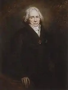 Portrait tout en contrastes d'un homme debout dans l'ombre, visible depuis la taille. Tout le tableau est sombre, marron et noir, excepté le visage. On distingue cependant un manteau noir et une main posée sur le bras d'un fauteuil marron. Le visage, figé, surmonte un col en V qui laisse voire une chemise blanche. Le regard est dur, les cheveux mi-long, bouclés et blancs.