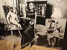 Photographie d'une femme en robe longue assise sur un tabouret, tenant une palette devant un nu posé sur un chevalet.