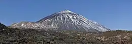Photo du volcan conique partiellement enneigé avec un dôme marqué à sa gauche et un terrain rocheux au premier plan.