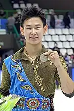 Un homme asiatique souriant en costume brun, vert et bleu, avec une médaille.