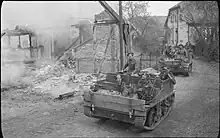 Photo noir et blanc de deux petits véhicules à chenille transportant des soldats au milieu de maisons en ruine.