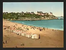 Vue panoramique d'une plage, avec des tentes pour abriter les baigneurs. Au fond une avancée rocheuse est surplombée de maisons.