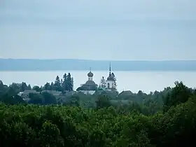 Photographie par temps couvert depuis les cimes d'une forêt d'églises et maisons d'un village bordant une grande étendue d'eau en arrière-plan.