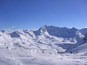 photographie en couleur montrant le domaine skiable de Tignes. Le lieu d'alpage pour les troupeaux l'été est ici, montré en hiver. Un télésiège en fonctionnement véhicule des sportifs au-dessus d'étendues enneigées en pentes douces. En arrière-plan, le relief très accentué montre la roche nue sur laquelle la neige ne peut s'accrocher.