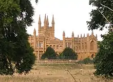 Photo d'un bâtiment en pierre ocre de grande taille, coiffé de nombreuses tourelles et caché derrière un rideau d'arbres
