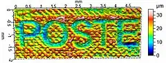 Micro-topographie d'un timbre-poste français d'usage courant (détail) montrant l'épaisseur d'encre obtenue par le procédé de taille-douce. Les caractères « POSTE » apparaissent sur le timbre en blanc sur fond rouge, et correspondent donc à une absence d'encre. La mesure a été réalisée à l'aide d'un profilomètre, et la fausse couleur représente l'altitude microscopique selon la légende figurant à droite.