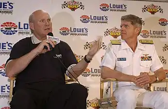 Un homme chauve habillé de noir tient un micro et discute sous les yeux d'un militaire paré de blanc.