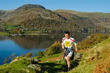 Coureur sur un sentier. En arrière-plan un lac et des collines vertes.