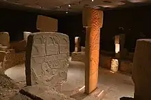 Deux pierres levées, chacune surmontées d'une autre pierre donnant une forme de T. À l'avant plan, il y a une pierre plate rectangulaire grise, posée sur une tranche et sculptée. Elle sont entourées de petits murs de pierres. Le tout est sous un plafond plat et illuminé par des projecteurs électriques.