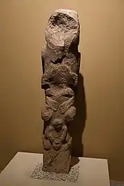 Pierre oblongue beige sculptée, posée sur une de ses petites faces carrées. Elle est posée sur un socle blanc face à un mur beige plus foncé. Le tout est éclairé par un projecteur électrique.