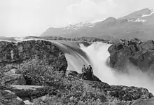 Photo en noir et blanc avec deux hommes assis devant une grosse chute d'eau, au milieu des montagnes.