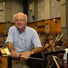 Un homme d'une soixantaine d'années posant en tournant le dos à un orchestre, la baguette de chef à la main, dans un studio d'enregistrement.