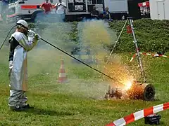 Un homme vêtu de manteaux protecteurs tient une longue tige d'où jaillit une boule de feu contre un bloc de métal.
