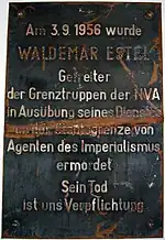 Plaque de métal endommagée indiquant « Am 3.9.1956 wurde WALDEMAR ESTEL Getreiter der Grenztruppen der NVA in Ausübung seine Dienstes an der Staatsgrenze von Agenten des Imperialismus ermordet. Sein Tod ist uns Verpflichtung. »