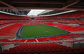 The interior of an empty stadium as viewed from its upper tier of seating. The seats are a vivid red and the pitch is a vivid green. The pale grey sky is visible through an opening in the ceiling above the pitch.