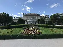 Des fleurs dessinent une horloge que des aiguilles parcourent. Derrière un grand bâtiment style demeure XIXème.