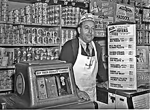 Photographie en noir et blanc d’un épicier souriant, cravaté, portant un tablier blanc à bretelle unique passant derrière le cou, coiffé d’un calot publicitaire ; l’homme s’appuie à une machine distributrice de café et à un comptoir surmonté de panneaux publicitaire ou annonçant les prix de marchandises. Derrière lui, l’étagère qui court tout le long du mur est remplie d’une multitude de boîtes de conserve, parfois cachées par des calicots publicitaires supplémentaires, donnant l’impression qu’il n’y a plus 1 cm2 de disponible.
